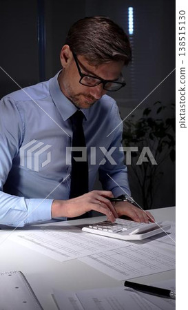 Accountant working late night in office, calculating finance data and economy statistics in a blue shirt with a tie. Audit and taxes in business Accountant working late night in office, calculating finance data and economy statistics in a blue shirt with a tie. Audit and taxes in business 138515130
