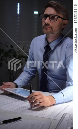 Businessman working late analyzing financial data at office, holding tablet with charts, writing notes on documents. Audit and taxes in business Businessman working late analyzing financial data at office, holding tablet with charts, writing notes on documents. Audit and taxes in business 138515146