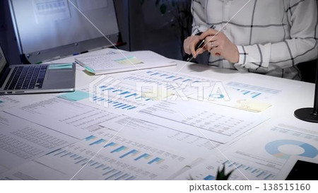 Financial advisor examining business reports and charts, analyzing data, planning investments, and preparing forecasts on a cluttered office desk in a late night work environment 138515160