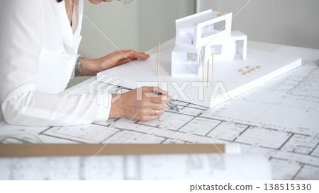 Female architect engrossed in architectural plans and miniature scale model, thoughtfully designing a new building project for modern construction development 138515330