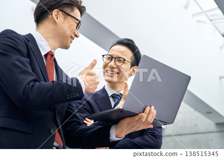 一張商務照片，照片中兩位日本商人面帶微笑，豎起大拇指，看著他們的電腦。 138515345