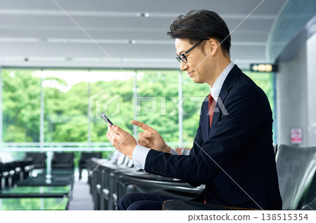 A smiling Japanese businessman operating his smartphone in an office lounge. A smiling Japanese businessman operating his smartphone in an office lounge. 138515354