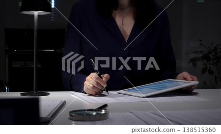Woman working late night in office, multitasking with a tablet and writing on documents, managing heavy paperwork load. Audit and taxes in business Woman working late night in office, multitasking with a tablet and writing on documents, managing heavy paperwork load. Audit and taxes in business 138515360