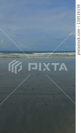 Vertical composition of a peaceful, deserted tropical beach showing calm ocean waves washing onto smooth, wet sand under an overcast sky. 138516156