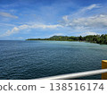Scenic coastal view of the Andaman sea from a pier, showing calm blue water and a lush, tree-lined tropical shore under a partly cloudy sky. 138516174