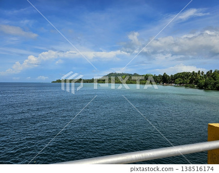 Scenic coastal view of the Andaman sea from a pier, showing calm blue water and a lush, tree-lined tropical shore under a partly cloudy sky. 138516174