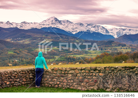 View of pastures in the valley and mountain peaks covered with first snow during sunset. Young woman looking at beautiful mountains. Viewpoint near Chapelle Notre-Dame-de-Roume. Cieutat, France View of pastures in the valley and mountain peaks covered with first snow during sunset. Young woman looking at beautiful mountains. Viewpoint near Chapelle Notre-Dame-de-Roume. Cieutat, France 138516646