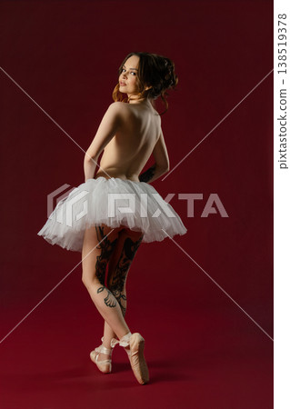 Elegant tattooed ballerina in a white tutu and pointe shoes looking back over her shoulder against a red background. Elegant tattooed ballerina in a white tutu and pointe shoes looking back over her shoulder against a red background. 138519378