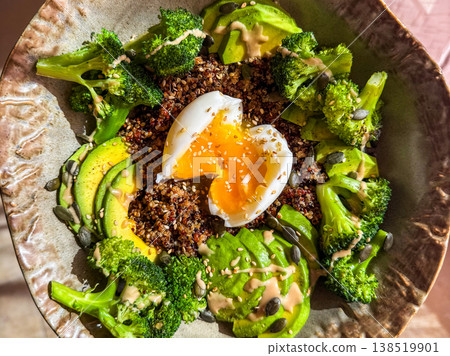 A healthy bowl featuring quinoa, soft-boiled egg, steamed broccoli, and sliced avocado. The dish is arranged in a textured bowl, showcasing vibrant colors and textures. 138519901
