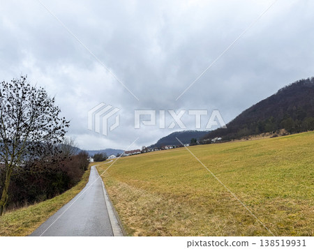 A scenic view of a rural landscape in Geislingen, Germany. A winding road leads through a grassy field under a cloudy sky, surrounded by hills. 138519931