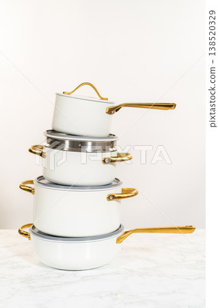 A vertical composition of four stacked ceramic pots in matte white finish, each with golden handles and matching lids. Photographed against a neutral background on a marble surface. Great for A vertical composition of four stacked ceramic pots in matte white finish, each with golden handles and matching lids. Photographed against a neutral background on a marble surface. Great for 138520329