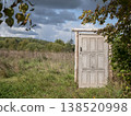 Freestanding weathered wooden door stands alone at the edge of an open field. The surreal scene evokes themes of escape, transition, and unexpected possibilities 138520998