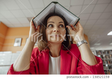 Close up young Caucasian woman with brown hair sits at a modern coworking office. She smiles while holding a book over her head with humor. The background features storage units and plants. 138521159