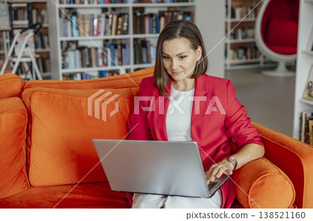 surprised woman working on laptop sitting on bright orange sofa in modern library interior, remote work education business coaching and digital communication 138521160
