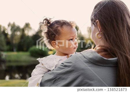 Mother Holding Toddler Daughter Outdoors at Sunset. Beautiful little child girl in arms of mom in green park 138521873