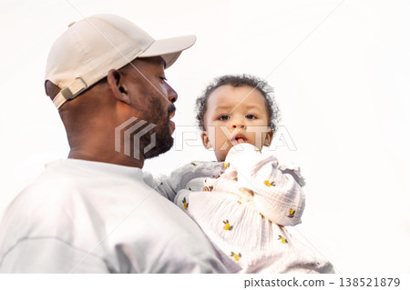 African American father holding beautiful little girl child with finger in mouth in arms Isolated against bright white sky background. Portrait dad with baby African American father holding beautiful little girl child with finger in mouth in arms Isolated against bright white sky background. Portrait dad with baby 138521879