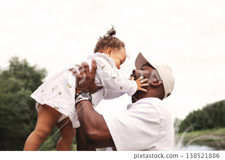Smiling African American father gently embracing toddler daughter against sky outdoors, loving hug, portrait with copy space. Happy dad with little child Smiling African American father gently embracing toddler daughter against sky outdoors, loving hug, portrait with copy space. Happy dad with little child 138521886