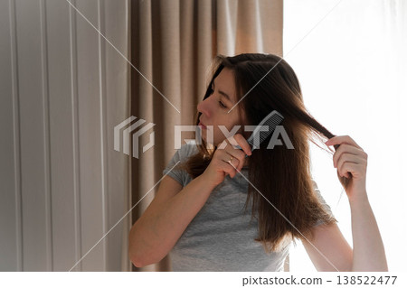 A woman combs her hair in front of a mirror. She's tidying herself up. She's wearing a casual gray T-shirt. Her expression is focused, as she prepares for the workday. Self-care. Space for text. A woman combs her hair in front of a mirror. She's tidying herself up. She's wearing a casual gray T-shirt. Her expression is focused, as she prepares for the workday. Self-care. Space for text. 138522477