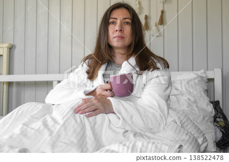 A woman lies on a pastel-colored bed, holding a coffee mug. A calm woman in a robe enjoys the serene morning atmosphere at home. Morning routine. Self-care. Starting a new day. Space for text. Banner 138522478
