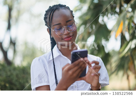 Young Black woman wearing glasses and a white shirt using smartphone outdoors, smiling while texting in a park setting. Mobile communication, social media, online lifestyle and technology concept. 138522566