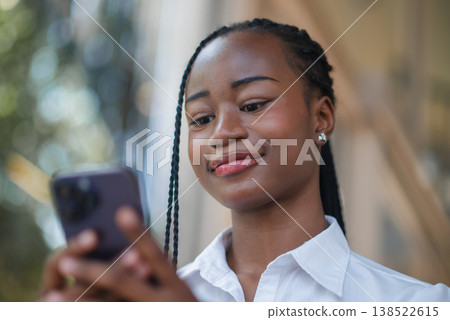 Low angle portrait of young Black woman looking at smartphone with a confident smile. Mobile app use, online connection, digital lifestyle, communication and modern technology concept. Low angle portrait of young Black woman looking at smartphone with a confident smile. Mobile app use, online connection, digital lifestyle, communication and modern technology concept. 138522615