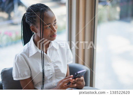 Young Black woman holding smartphone while sitting by a window, looking away thoughtfully. Modern mobile lifestyle, waiting moment, digital connection, communication and indoor calm. Young Black woman holding smartphone while sitting by a window, looking away thoughtfully. Modern mobile lifestyle, waiting moment, digital connection, communication and indoor calm. 138522620