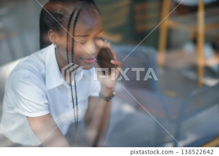 Young Black woman speaking on smartphone behind cafe window, seen through glass reflections. Candid business lifestyle, remote conversation, modern communication and indoor connection. Young Black woman speaking on smartphone behind cafe window, seen through glass reflections. Candid business lifestyle, remote conversation, modern communication and indoor connection. 138522642