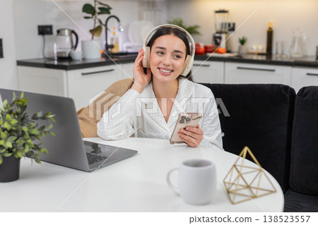 Young woman wears wireless headphones types message on smartphone takes break with laptop nearby Young woman wears wireless headphones types message on smartphone takes break with laptop nearby 138523557