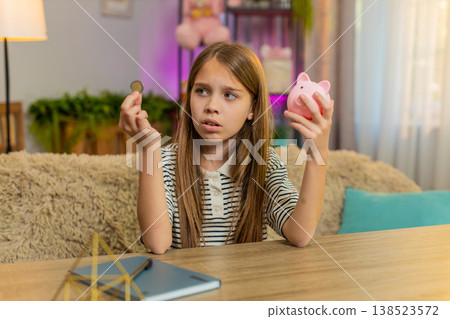 Child girl kid sadly looks at empty piggy bank shaking head at home table feeling disappointment 138523572