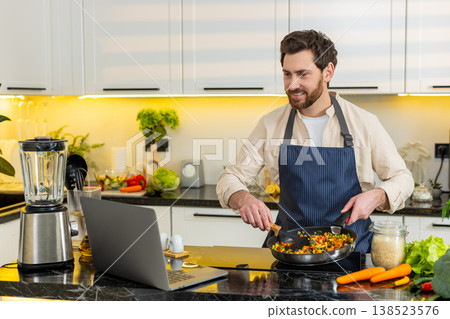 Middle-aged man cooking vegetables on electric stove while watching laptop recipe for healthy diet Middle-aged man cooking vegetables on electric stove while watching laptop recipe for healthy diet 138523576