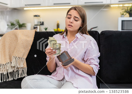 Young woman at home looks inside wallet sees small cash money dollars frowns about poverty debt Young woman at home looks inside wallet sees small cash money dollars frowns about poverty debt 138523586