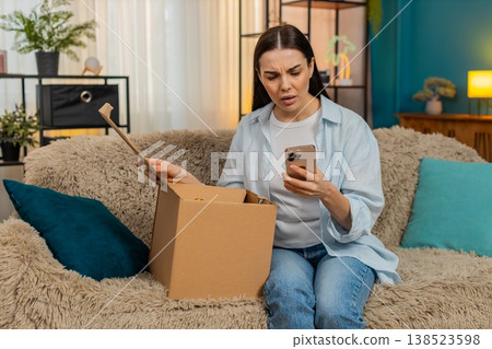Young woman sitting on sofa unpacking delivery finding wrong item disappointed shaking head sadly Young woman sitting on sofa unpacking delivery finding wrong item disappointed shaking head sadly 138523598