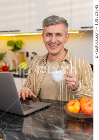 Middle-aged man working on laptop with coffee in kitchen for freelance work and remote business task 138523642