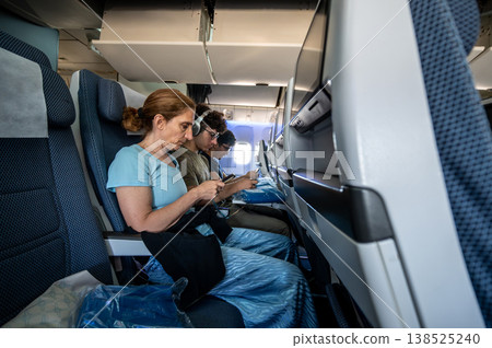 Passengers are sitting in economy class airplane seats, using their personal smartphones and wearing headphones, engrossed in digital entertainment and communication during a flight Passengers are sitting in economy class airplane seats, using their personal smartphones and wearing headphones, engrossed in digital entertainment and communication during a flight 138525240