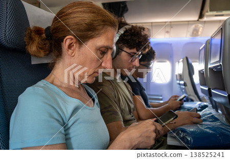 Passengers seated in an airplane cabin focus on their mobile devices during a flight, showing modern technology use and digital connectivity while traveling together Passengers seated in an airplane cabin focus on their mobile devices during a flight, showing modern technology use and digital connectivity while traveling together 138525241