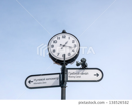 Large round clock on Ob River embankment with signs Rowing Canal and Zero Kilometer against sky covered in snow cold early spring urban landscape. 138526582