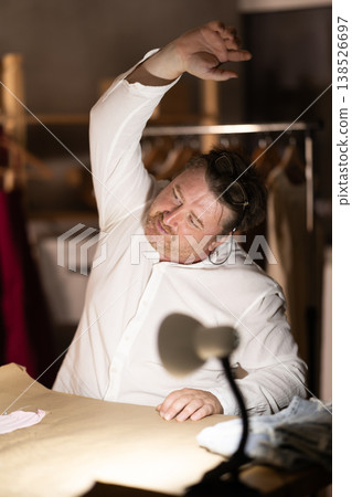 Middle-aged male tailor stretching while sitting next to sewing machine at night Middle-aged male tailor stretching while sitting next to sewing machine at night 138526697