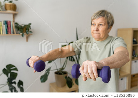 An elderly woman does arm exercises with dumbbells at home, maintaining fitness and strength, promoting wellness, and healthy aging. Mindful happiness, an active, and healthy lifestyle An elderly woman does arm exercises with dumbbells at home, maintaining fitness and strength, promoting wellness, and healthy aging. Mindful happiness, an active, and healthy lifestyle 138526896