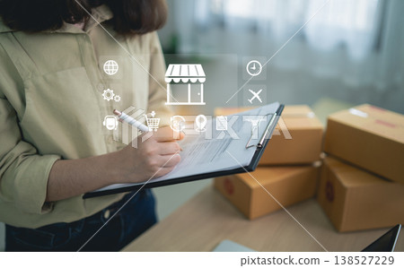 Person Writing on Clipboard with Digital E-commerce Icons in Modern Workspace Surrounded by Cardboard Boxes and Shipping Supplies Person Writing on Clipboard with Digital E-commerce Icons in Modern Workspace Surrounded by Cardboard Boxes and Shipping Supplies 138527229