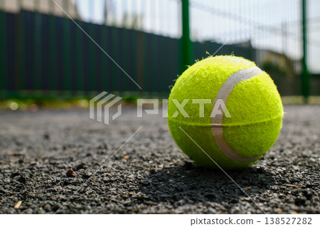 Yellow tennis ball resting on gray asphalt court surface with green fence background in outdoor sports area Yellow tennis ball resting on gray asphalt court surface with green fence background in outdoor sports area 138527282