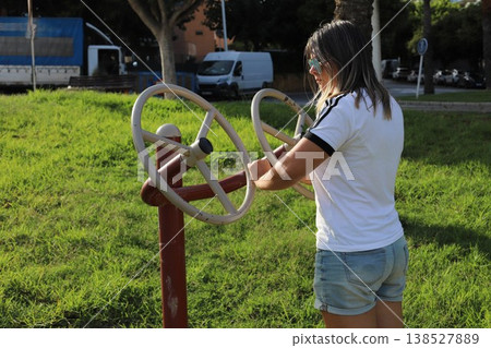 Young woman exercises on outdoor fitness equipment in a park, surrounded by green grass and trees, with vehicles visible in the background Young woman exercises on outdoor fitness equipment in a park, surrounded by green grass and trees, with vehicles visible in the background 138527889