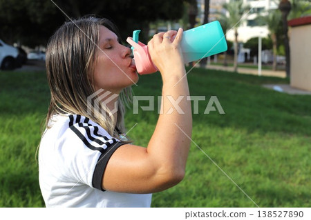 Woman with long hair drinks water from a colorful sports bottle while standing in a green park on a sunny day 138527890
