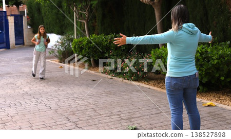 Mother wearing a light blue sweater stands with open arms on a paved pathway, welcoming dougher  138527898