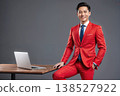 Confident Asian man in a vibrant red suit posing beside a laptop on a wooden table 138527922