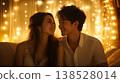 Smiling couple sitting close on a couch with warm string lights in the background, looking at each other lovingly 138528014