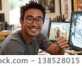 Smiling man with glasses giving thumbs up at desk with computer showing cartoon drawing in studio with art supplies in background 138528015