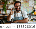 Smiling bearded man with glasses in apron gives thumbs up while leaning on desk with paper map in cozy workspace 138528016