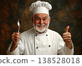 Smiling senior chef in white uniform and hat holding a spoon and giving a thumbs up on a dark studio background 138528018