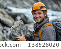 Smiling climber in orange helmet holding a rock, wearing backpack and jacket in snowy mountain setting 138528020