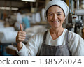 Smiling female worker in apron and headscarf giving thumbs up in a factory interior 138528024